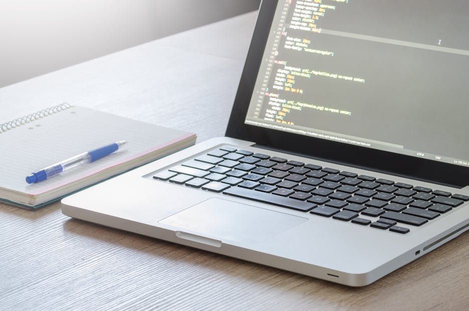 A close-up view of a laptop with an open screen displaying lines of colorful code, placed on a light wooden desk. To the left of the laptop, there is a spiral-bound notebook with a blue pen resting on