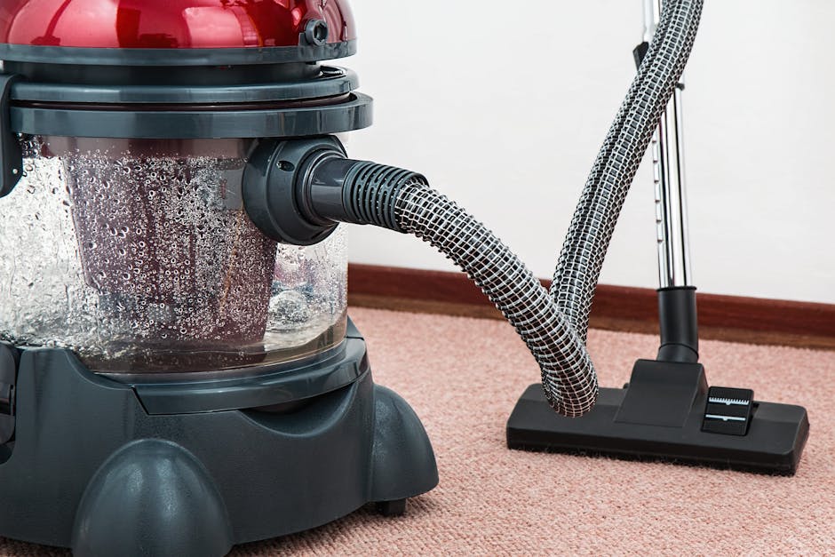 Close-up of a vacuum cleaner with a transparent dust container and a flexible, patterned hose connected to a floor cleaning head on a pinkish carpeted surface, indoors with plain white walls and woode