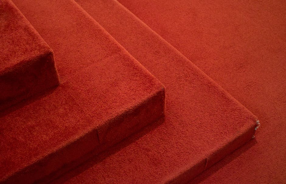Close-up of a vacuum cleaner with a transparent dust container and a flexible, patterned hose connected to a floor cleaning head on a pinkish carpeted surface, indoors with plain white walls and woode